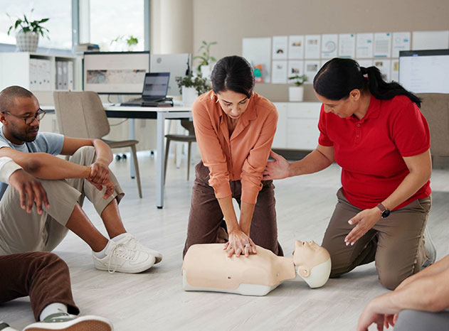 Dos mujeres practican maniobras de reanimación cardiopulmonar sobre un maniquí, con un hombre sentado observando